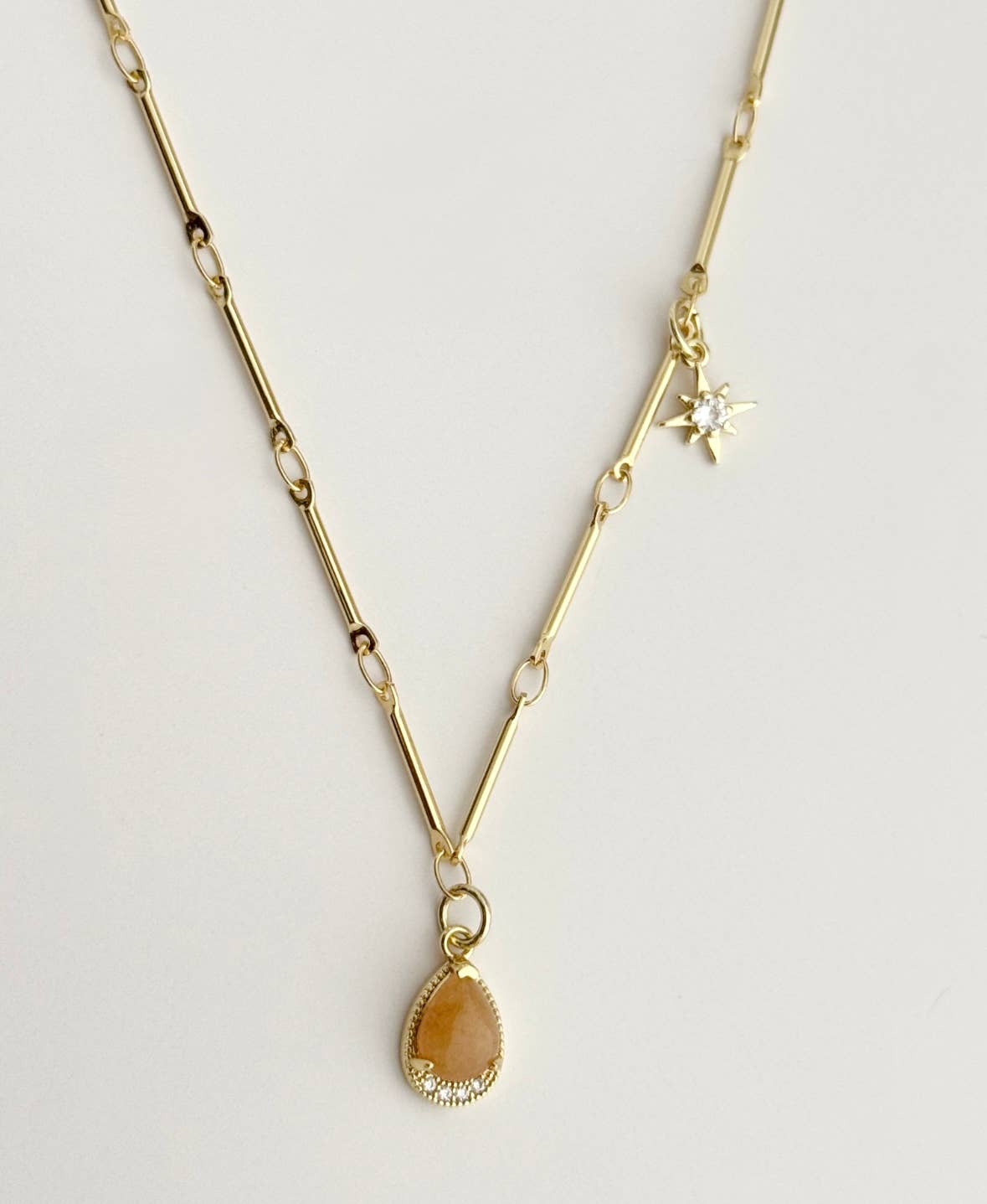 The Stone From The Sun Necklace by Tramps+Thieves features an 18K gold-filled elongated link chain, a star-shaped CZ charm, and a tan-centered teardrop pendant surrounded by crystals.