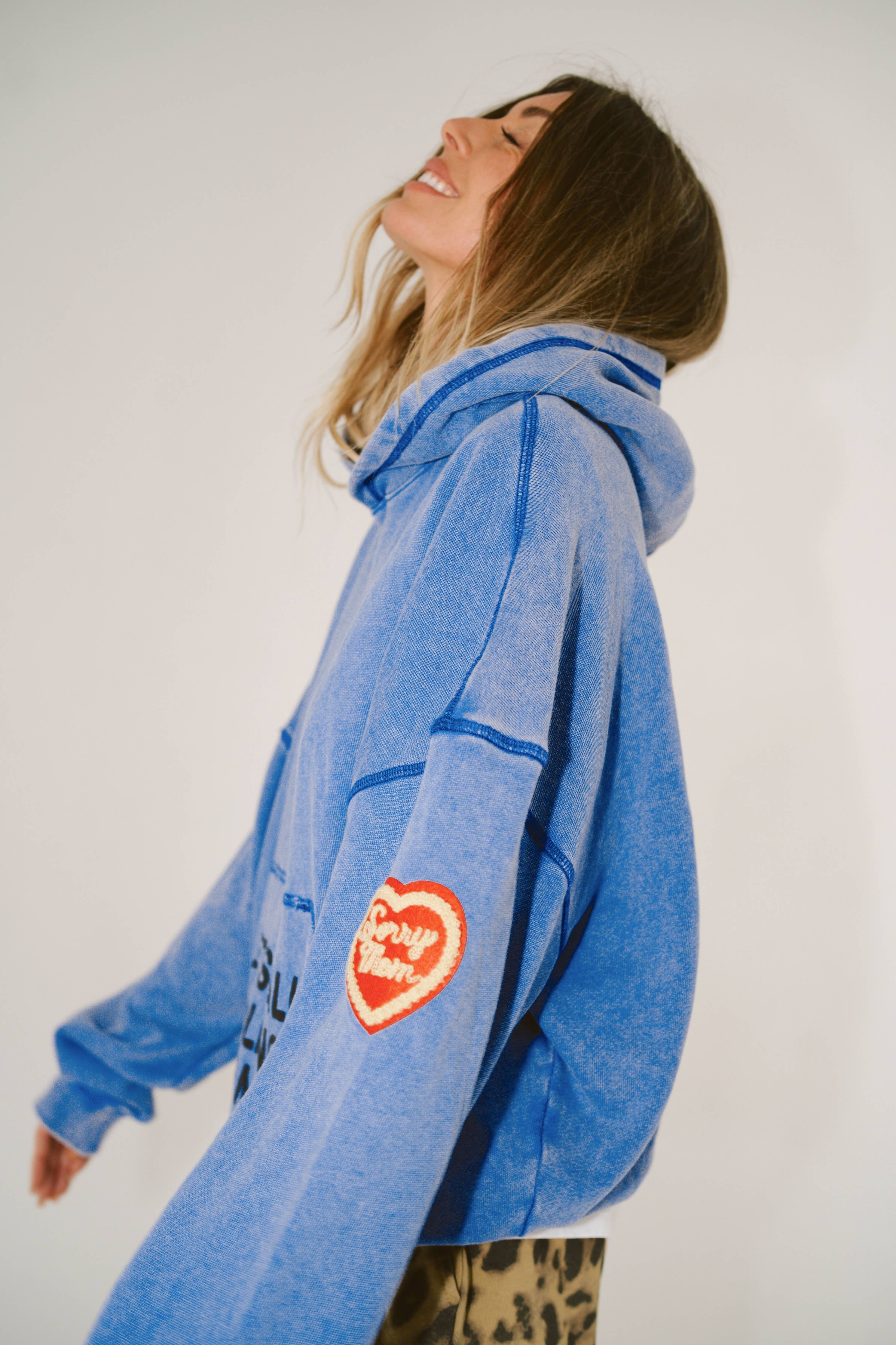 Woman wearing blue SORRY MOM HOODIE - PRESALE with heart patch on sleeve, smiling with eyes closed against plain background