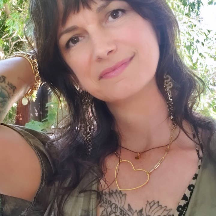 A woman with wavy brown hair smiles gently outdoors, wearing the Dreamers of Dreams Big Gold Heart | Paperclip Chain Necklace. Sunlight filters through green leaves, highlighting her large earrings and floral chest tattoo.