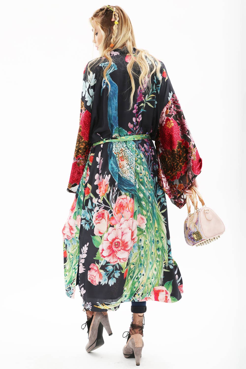 A woman with long blonde hair models the Aratta Sweet Fantasy Kimono, featuring burnout velvet sleeves and a peacock floral pattern. Facing away from the camera, she holds a pink handbag and stands in high heels against a white backdrop.