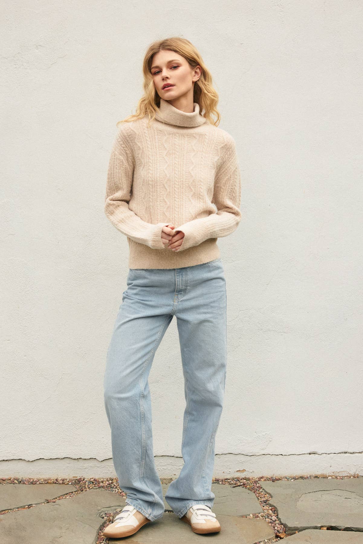 A person with long blonde hair stands against a plain light wall, wearing the Dress Forum Fluffy Cable Detail Turtle Neck Sweater in beige, light blue jeans, and white sneakers. Their hands are clasped in front and they look relaxed.