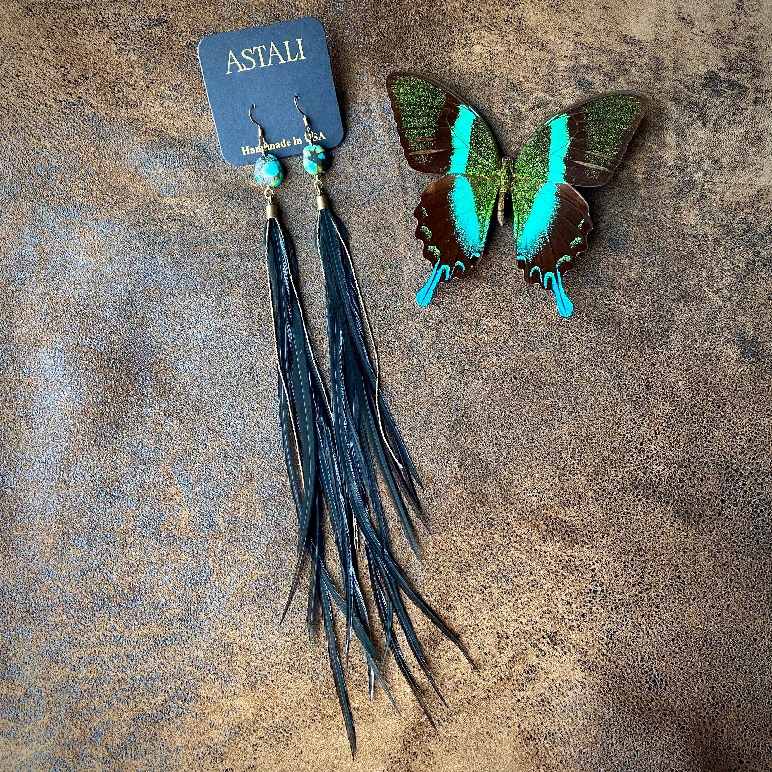 A pair of ASTALI Feather & Turquoise Statement Earrings, handcrafted in the USA from antique brass and sustainable coque feathers, rest beside a green and turquoise butterfly on a brown textured surface.