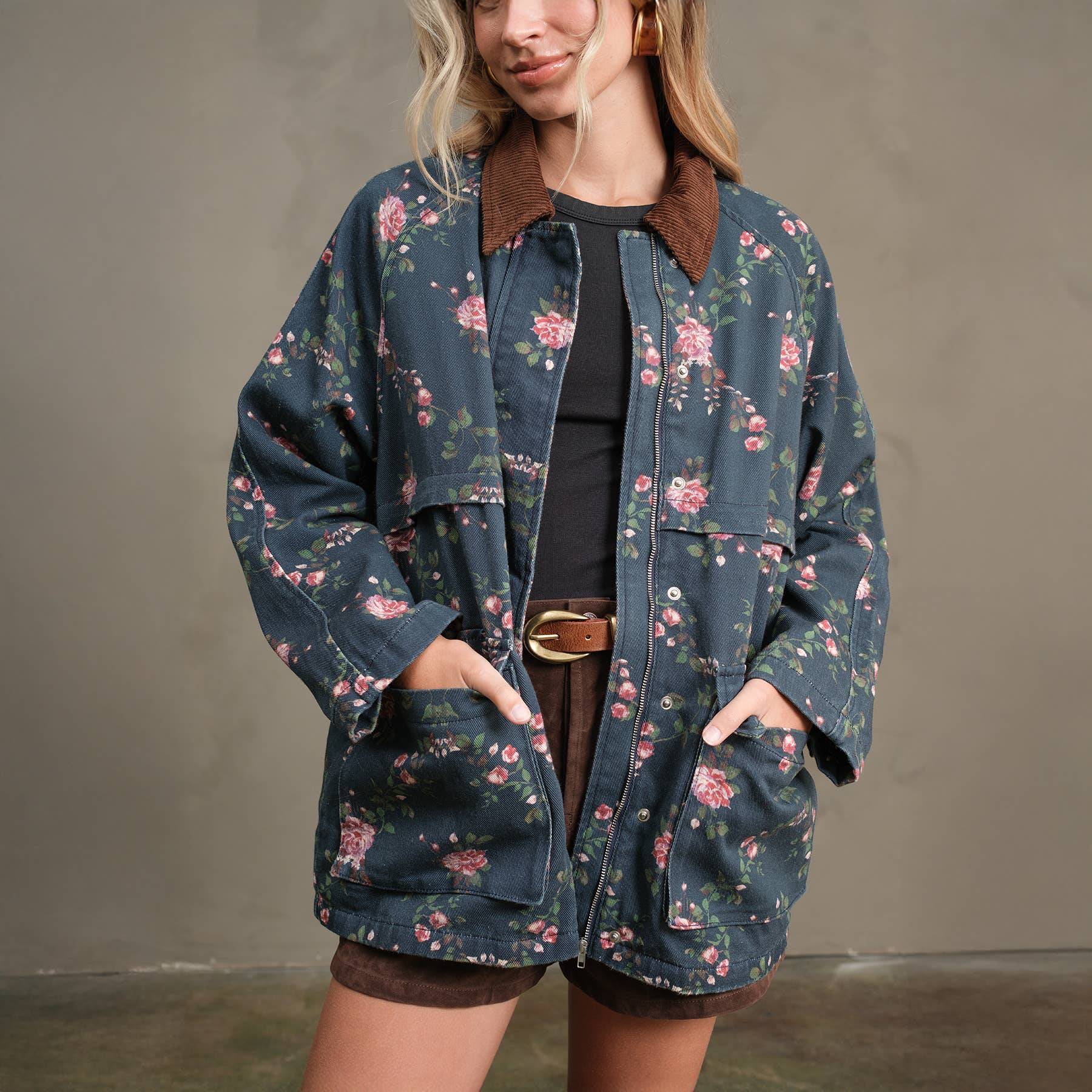 A woman wears the RAISONNEL Floral Printed Utility Jacket (RJ1073) with a brown collar and cuffs over a black top and brown shorts, standing hands-in-pockets against a neutral background.