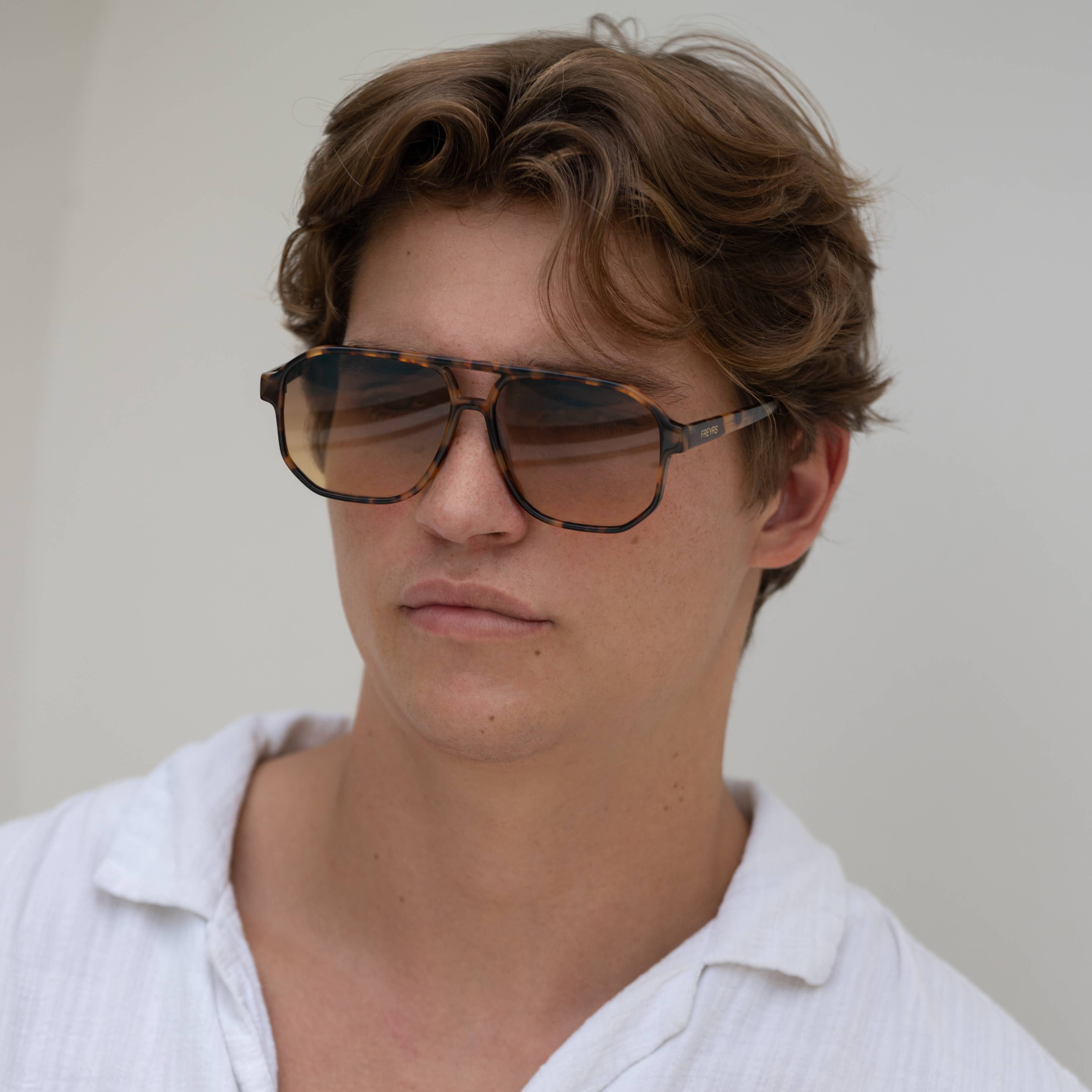 A young man with wavy brown hair wears FREYRS Eyewear Billie mens aviator sunglasses, crafted from biodegradable acetate, and a white collared shirt, gazing to the side against a plain light background.