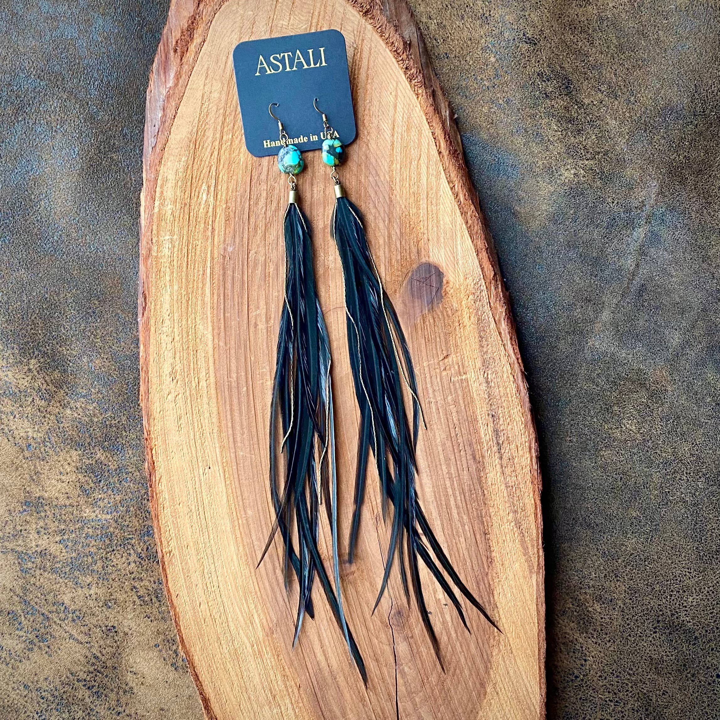 Feather & Turquoise Statement Earrings by ASTALI feature sustainable coque feathers, turquoise beads, and antique brass. Handmade in the USA, these bold black earrings are displayed on a branded card atop a wooden slab.
