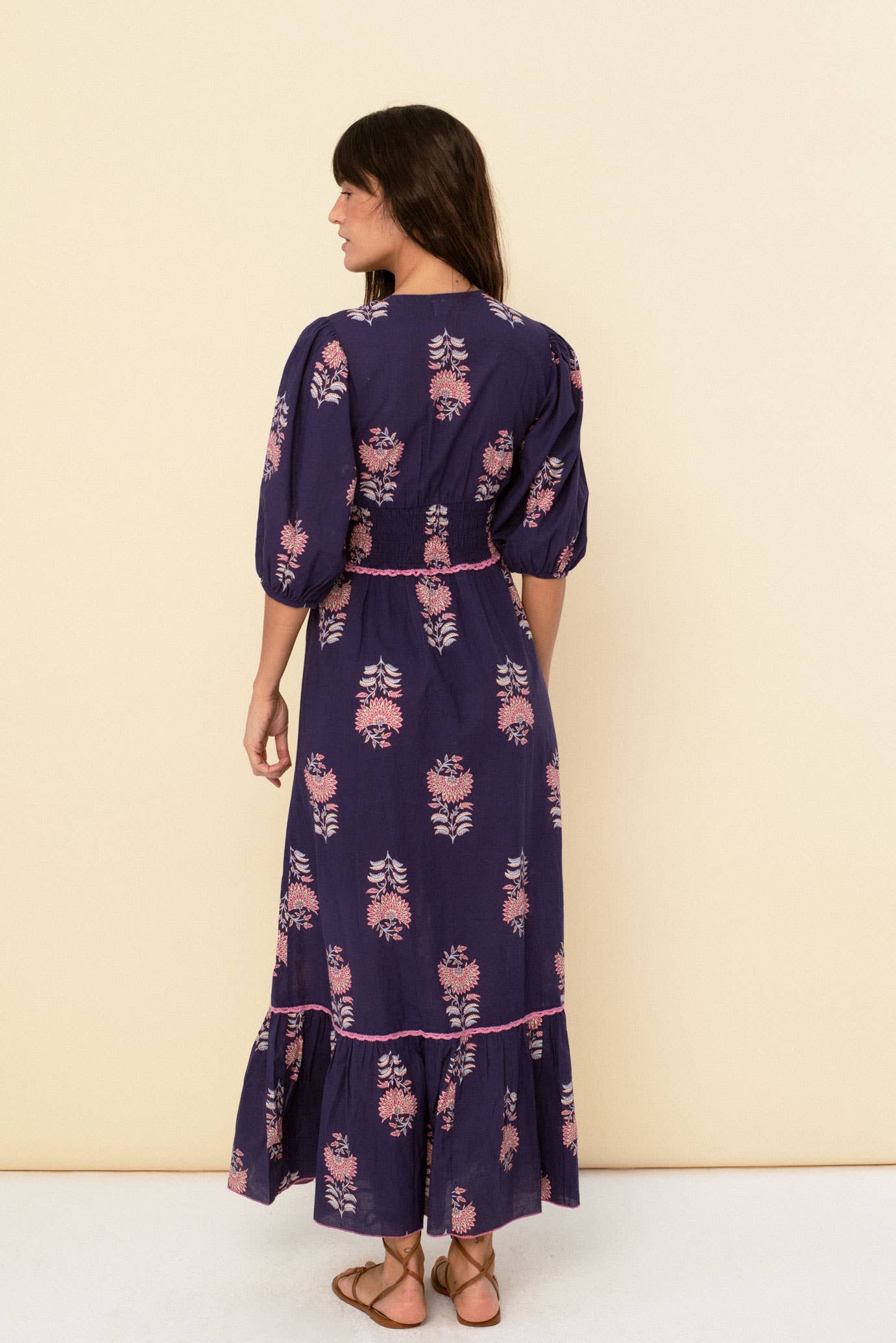 A woman with long brown hair faces away, wearing the Magnolia dress blue by Sonmer Shop USA—a romantic vintage navy maxi with pink florals, puffed half sleeves, and a ruffled hem. She pairs it with flat sandals against a light beige background.