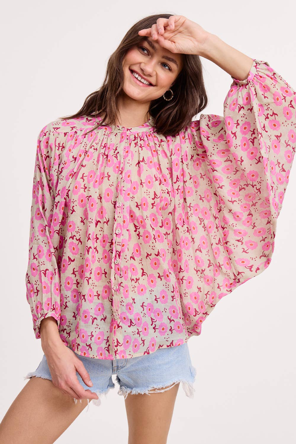 A smiling woman poses against a plain background, wearing the Ultramarine Meadow Breeze Linen Blouse—an airy, long-sleeved top in pink floral print—paired with light denim shorts and touching her forehead with one hand.