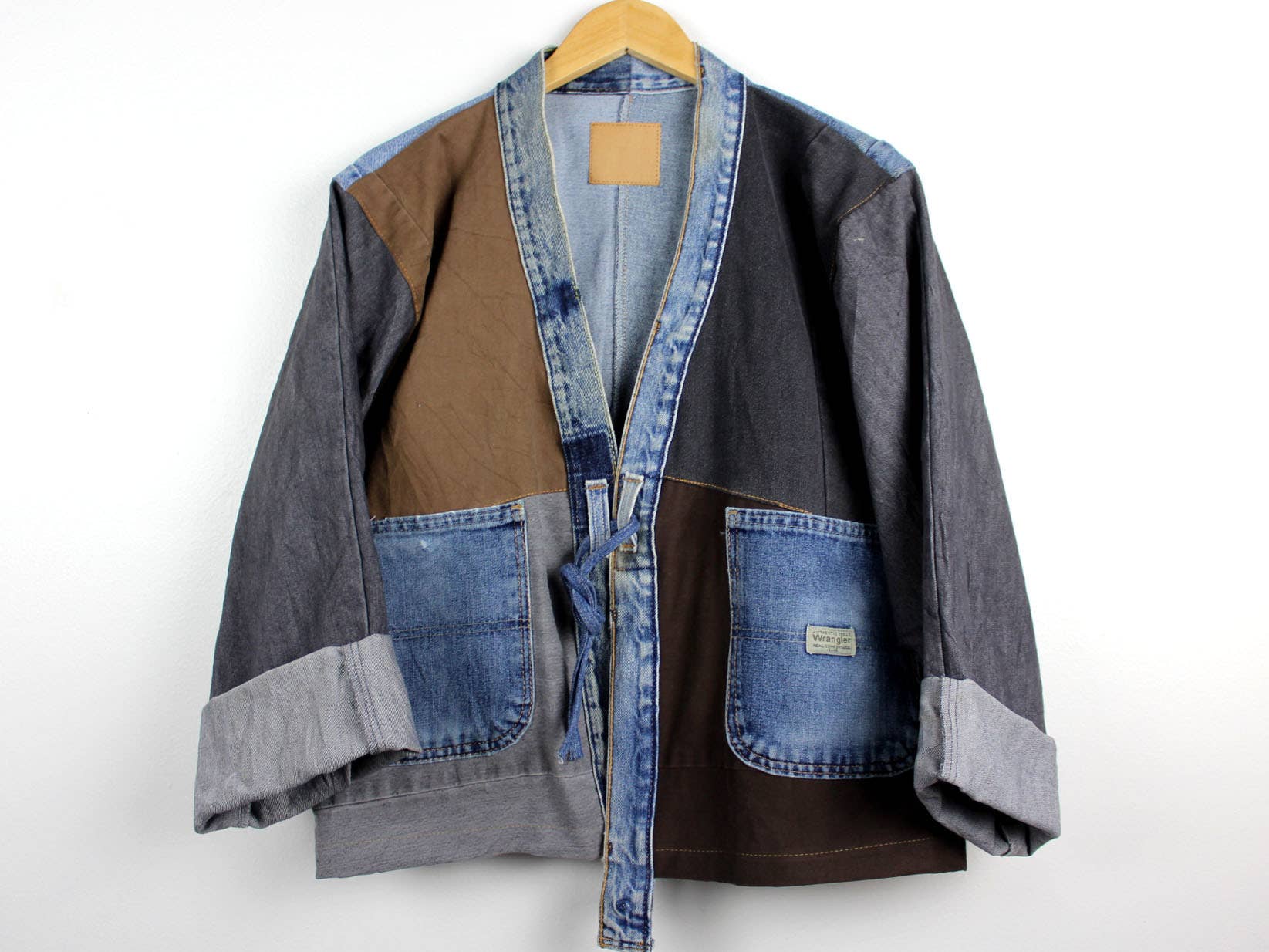 Upcycled Patchwork Denim Jacket with mixed denim and brown fabric patches, front pockets, and a relaxed fit on a hanger