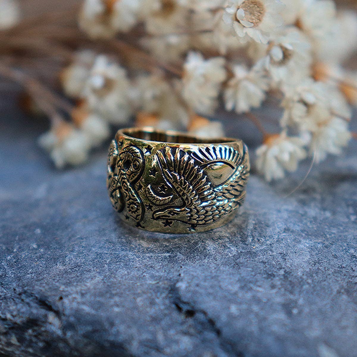 The Rainbow Serpent Ring by bahgsu jewels, a Xavier Rudd collaboration, is showcased on a stone surface with softly blurred dried flowers in the background.