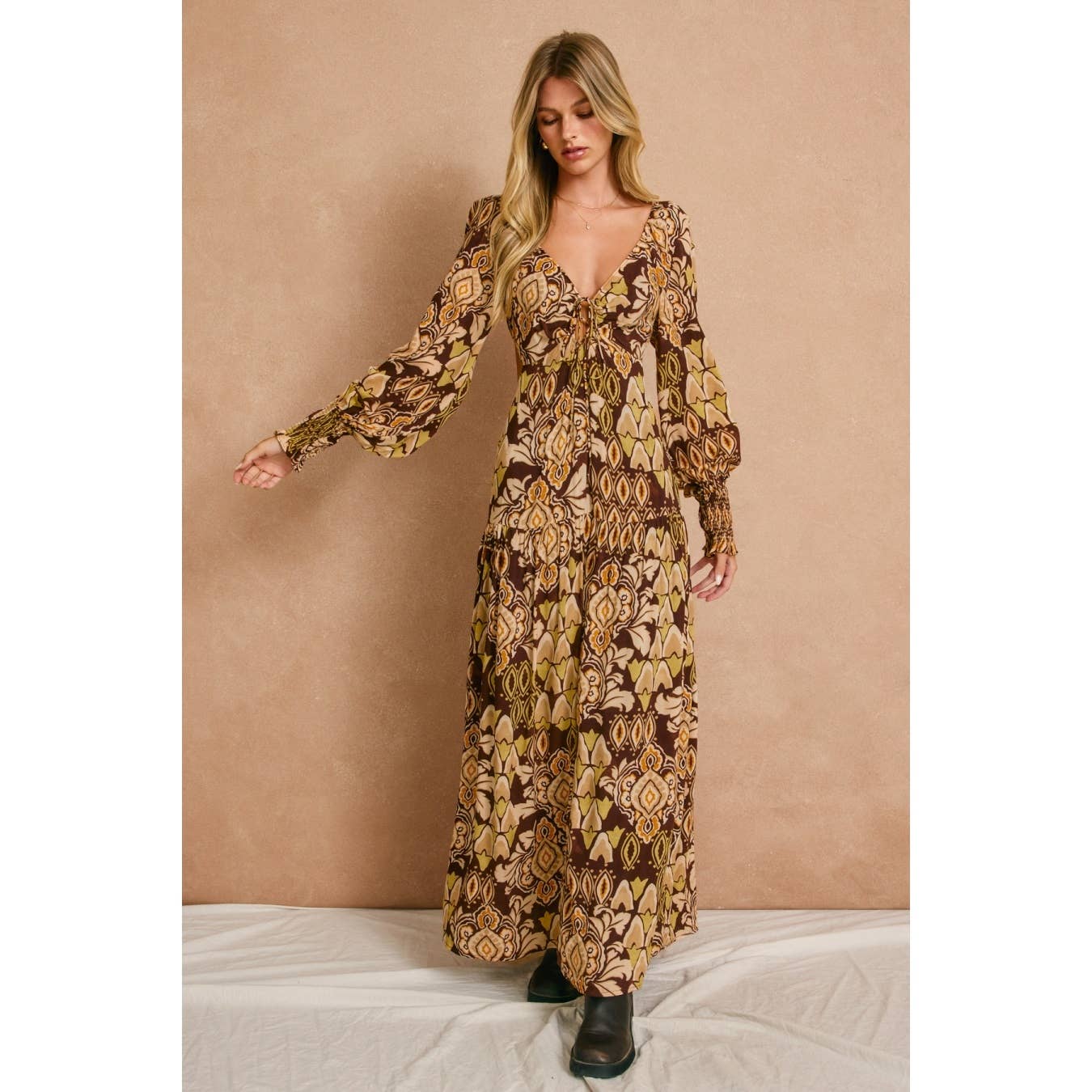 A woman in the Dress Forum Stone Ember Keyhole Long Sleeve Maxi Dress, featuring earth-toned geometric patterns, stands against a beige backdrop. She has long blonde hair and wears black shoes to complete her look.