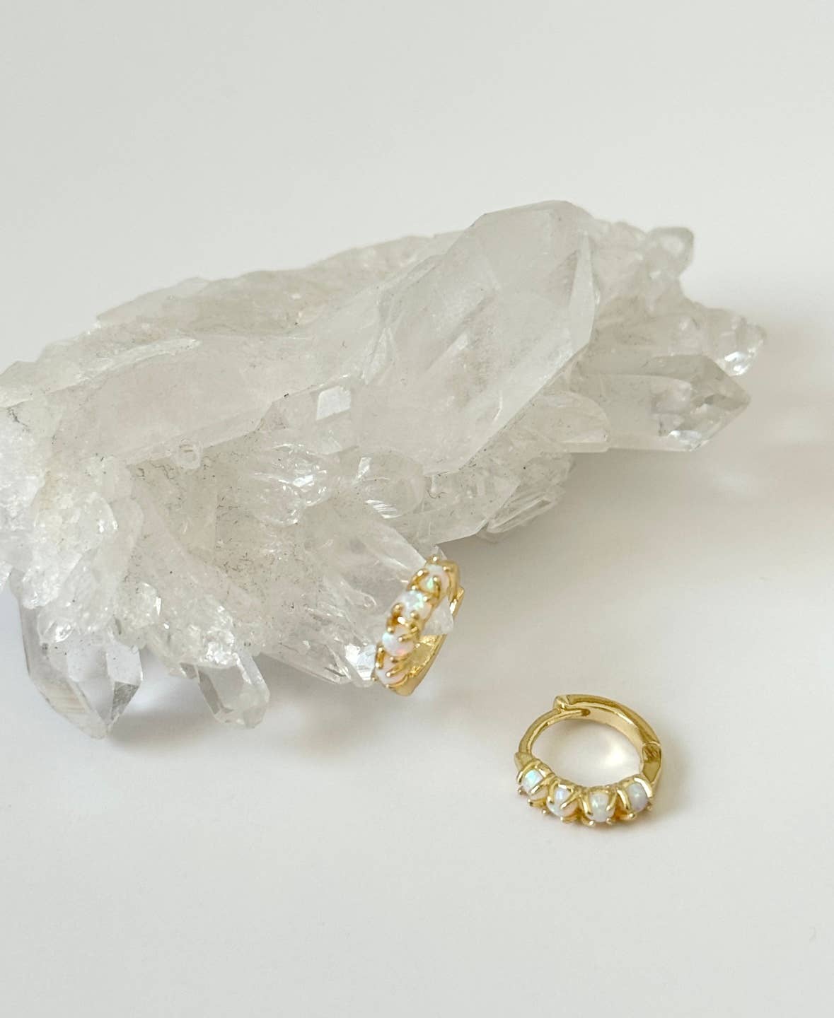 The Sonata Opal Hoops by Tramps+Thieves, gold-plated with white gemstone accents, are displayed—one on clear quartz and the other on a white surface—showcasing modern elegance in opal earring design.