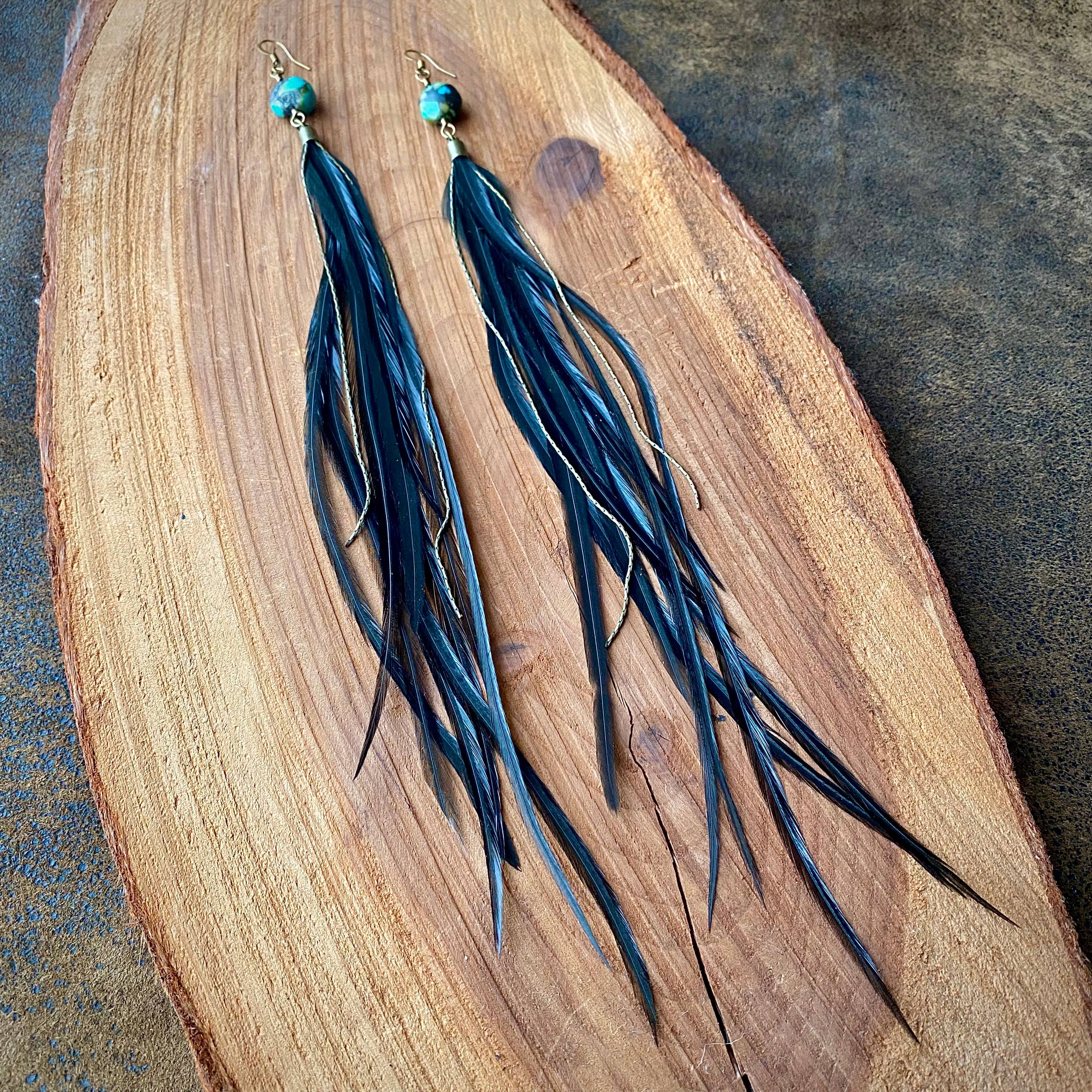ASTALIs Feather & Turquoise Statement Earrings feature sustainable coque feathers, turquoise bead accents, and antique brass. They have a natural, flowing look and are displayed on smooth wood.