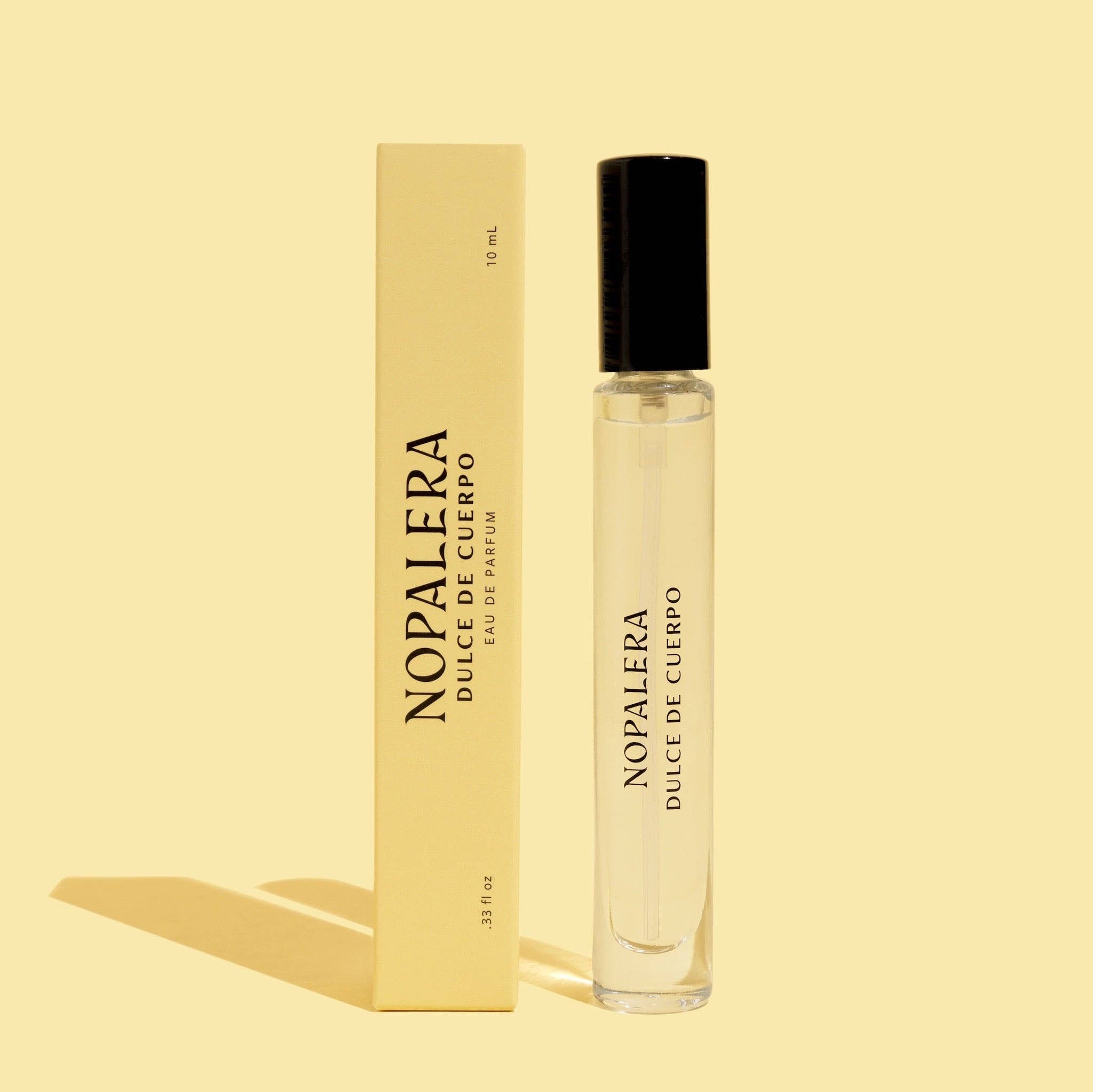 A clear glass Nopalera Dulce de Cuerpo Eau de Parfum – 10mL Travel Spray stands upright beside its matching beige box on a beige background. Both feature black text and highlight this gender-neutral sandalwood vanilla fragrance.