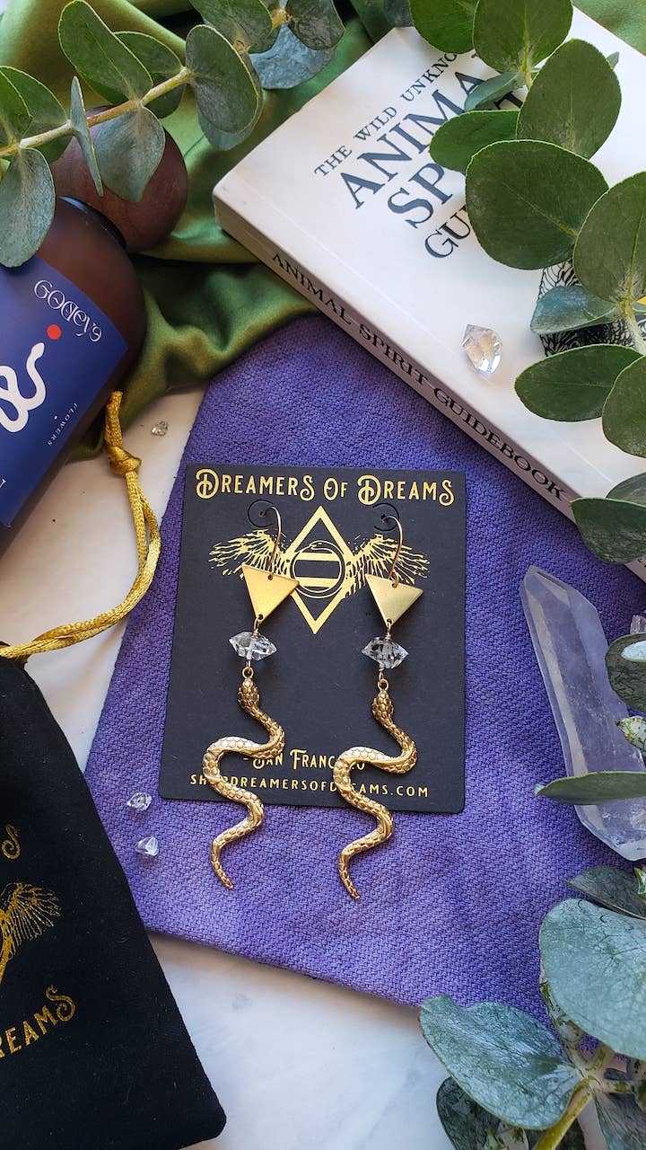 Gold Year of the Snake Earrings by Dreamers of Dreams are shown on a black and gold card, surrounded by eucalyptus leaves, crystals, a purple cloth, books, and a black pouch.