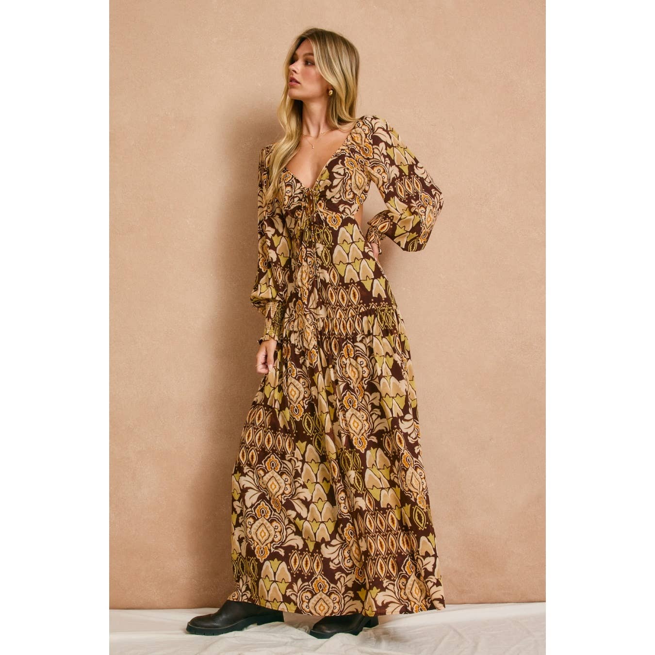 A woman with long blonde hair models the Dress Forum Stone Ember Keyhole Long Sleeve Maxi Dress in brown and yellow prints, standing against a light beige background and gazing to the side.