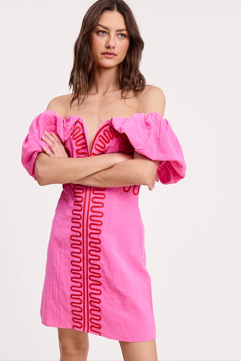 A woman stands with arms crossed, wearing the Ultramarine Embroidery Detail Off Shoulder Linen Mini Dress with voluminous ruffle sleeves and bold red embroidery. The background is plain and light-colored.