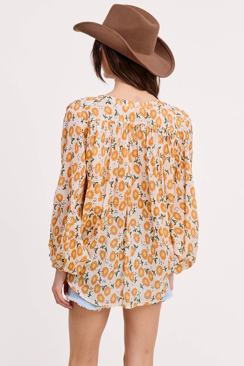 A woman seen from the back against a plain light background wears Ultramarine’s Meadow Breeze Linen Blouse, featuring yellow and orange floral prints, paired with a brown cowboy hat and light denim shorts.