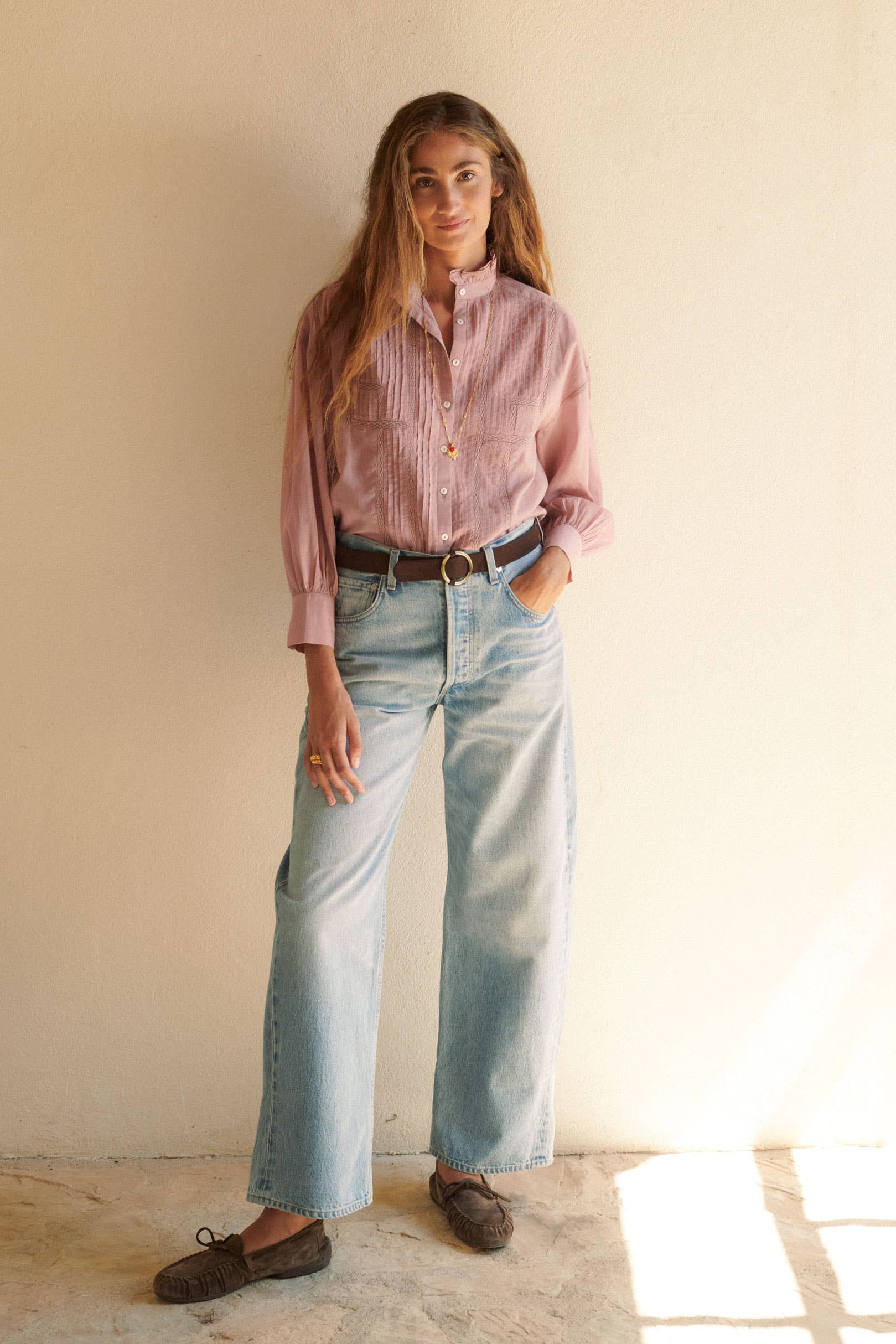 A woman stands against a light wall, wearing the Alexa Blouse Rose by Sonmer Shop USA with lace trim, paired with light blue wide-leg jeans, a brown belt, and loafers—her long wavy hair and relaxed pose make this look ideal for fall layering.
