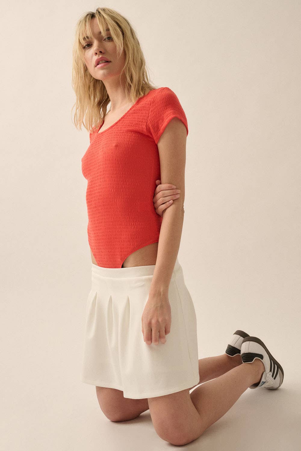Woman kneeling wearing a bright red Smocked Jersey Scoop Neck Bodysuit paired with a white skirt and sneakers