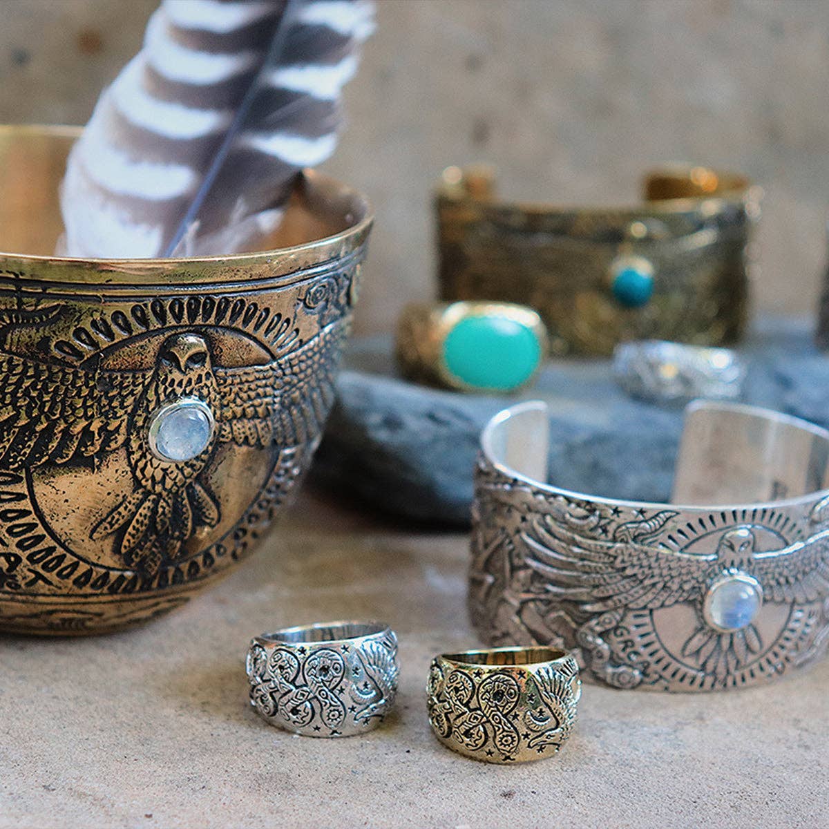The Rainbow Serpent Ring from bahgsu jewels Xavier Rudd Collab, featuring engraved bird designs and gemstones, is displayed with a decorative metal bowl, a feather, and a stone in the background.