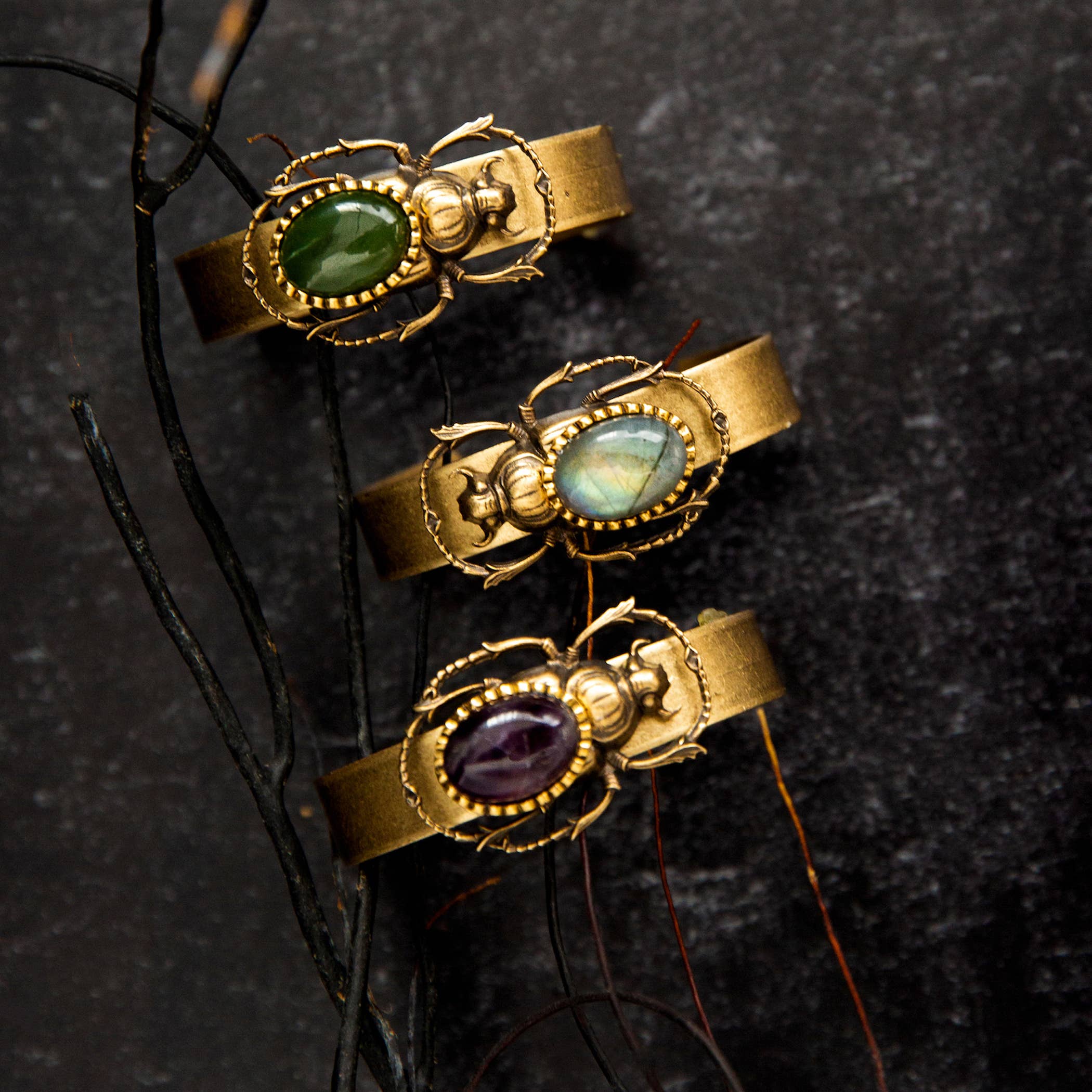 Three Sacred Scarab Cuff bracelets featuring green, blue, and purple stone scarab designs on brass bands.