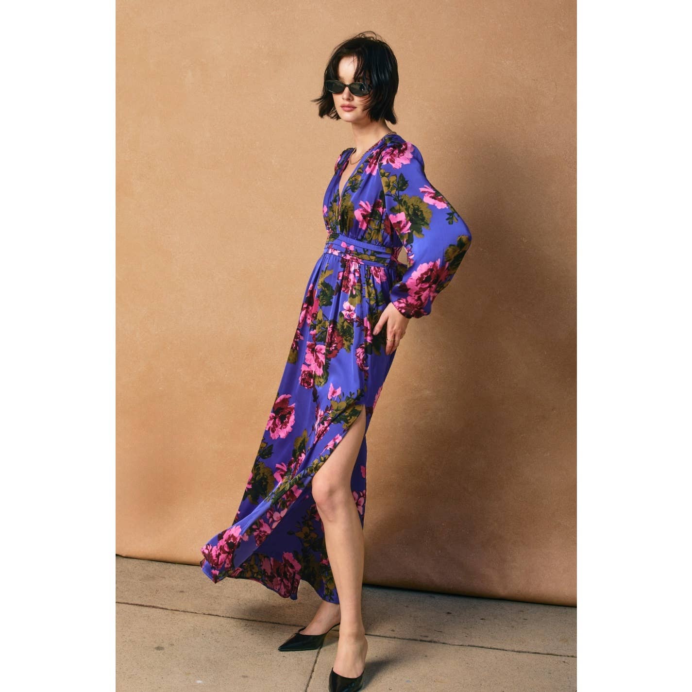 A woman poses confidently against a beige background in the Dress Forum Mesmerizing Essence Pleated Waist Maxi Dress, featuring a floral print, pleated waist, high side slit, paired with black heels and sunglasses.