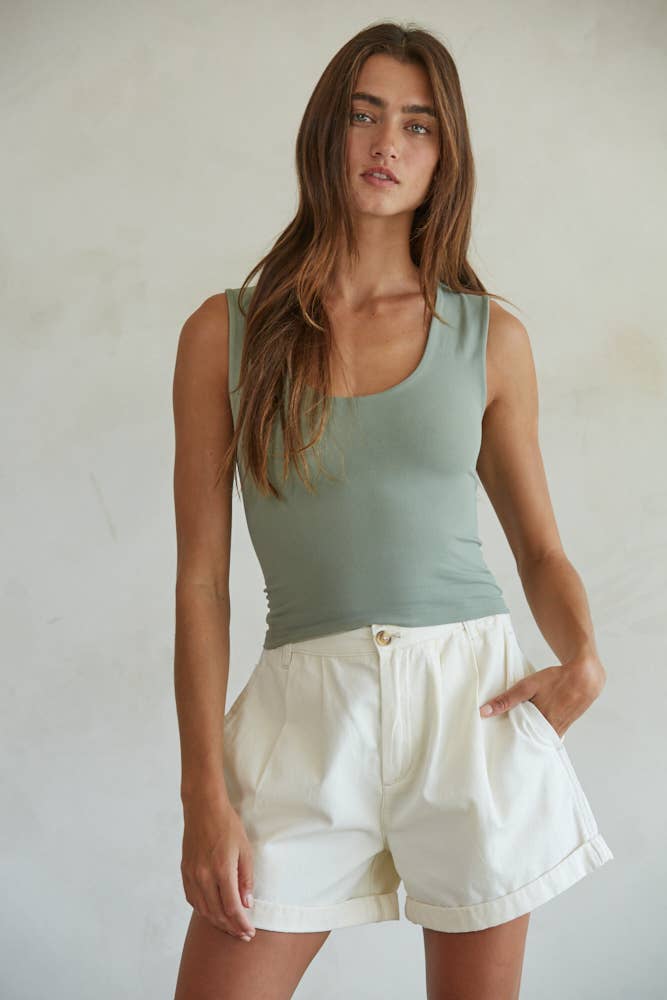 A woman with long brown hair wears the By Together Double Layered Seamless Top in sage green and high-waisted white shorts, standing against a light, plain background with one hand in her pocket.