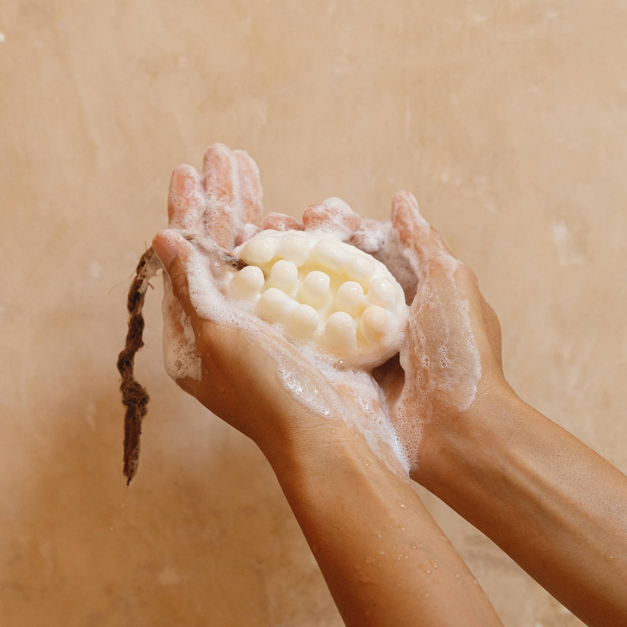 Two hands hold Cedar and Myrrhs Palo Santo + Shea Butter Bar Soap, a lathered, handmade bar shaped like bubbles with an attached rope—ideal for sensitive skin—set against a beige background.