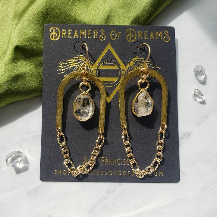 Gold + Glam Arch Chain Earrings by Dreamers of Dreams feature hammered brass hoops, clear faceted crystals, green fabric details, chain accents, and scattered crystals, all displayed on a sleek black branded card.