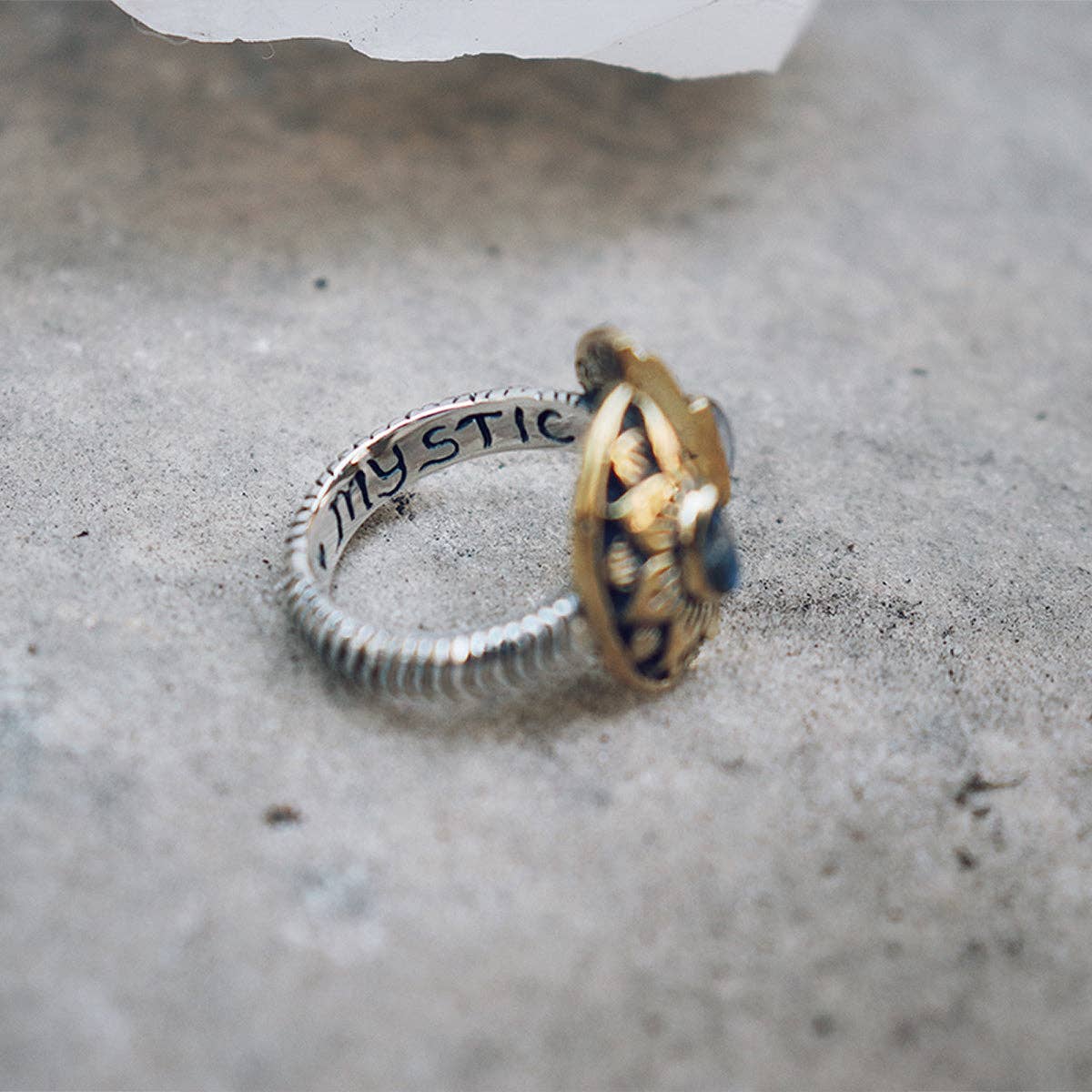 The Natural Mystic Ring - Moonstone by bahgsu jewels, crafted in sterling silver with MYSTIC engraved, features a gold-accented top and blue moonstone, displayed on a textured gray surface.