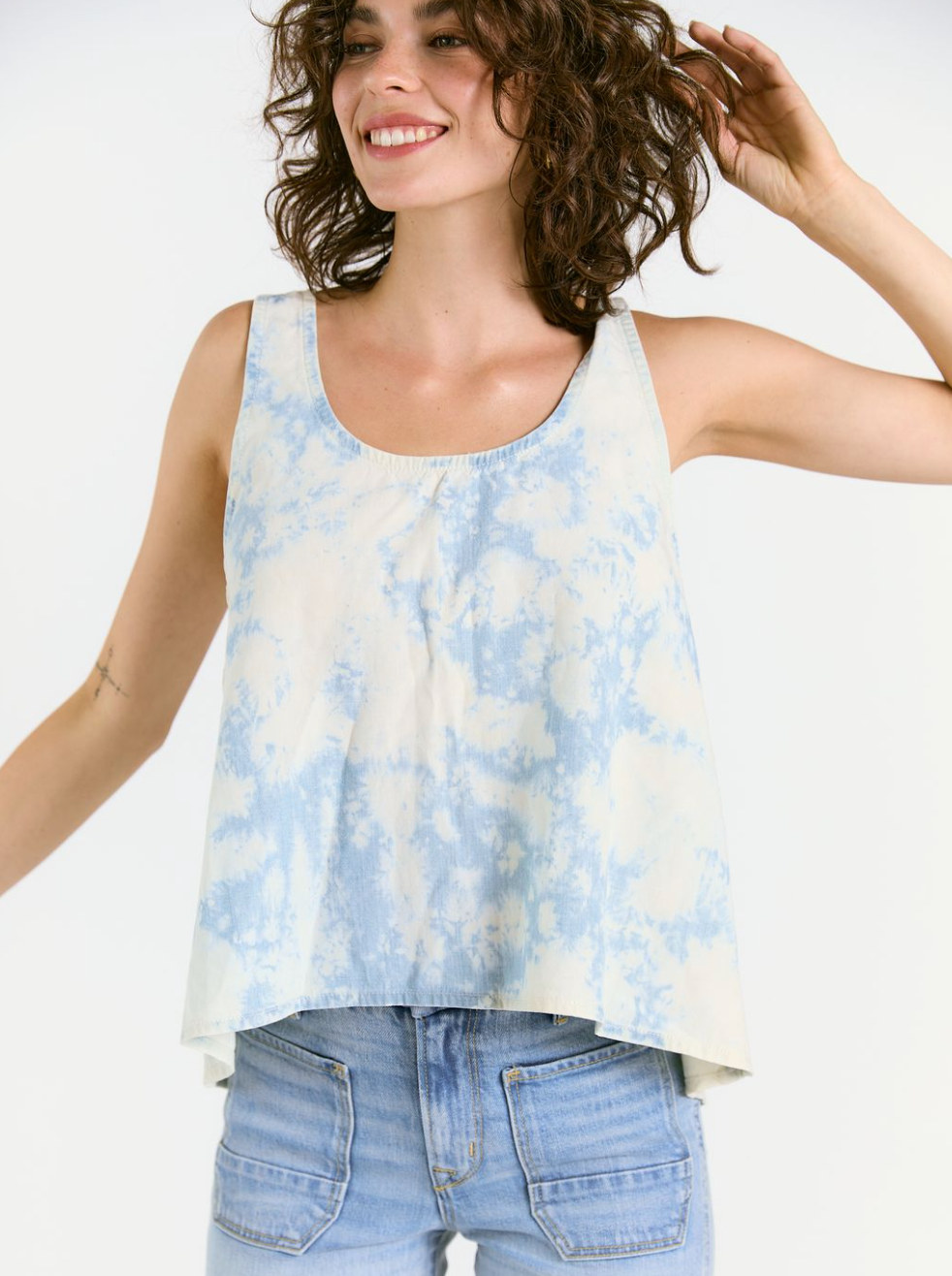 A smiling woman with curly brown hair models the Porter Blue Apparel Wabi Sabi Tank in Sky Tie Dye—a sleeveless, loose-fitting, eco-friendly top—paired with high-waisted light blue jeans, standing against a white background.