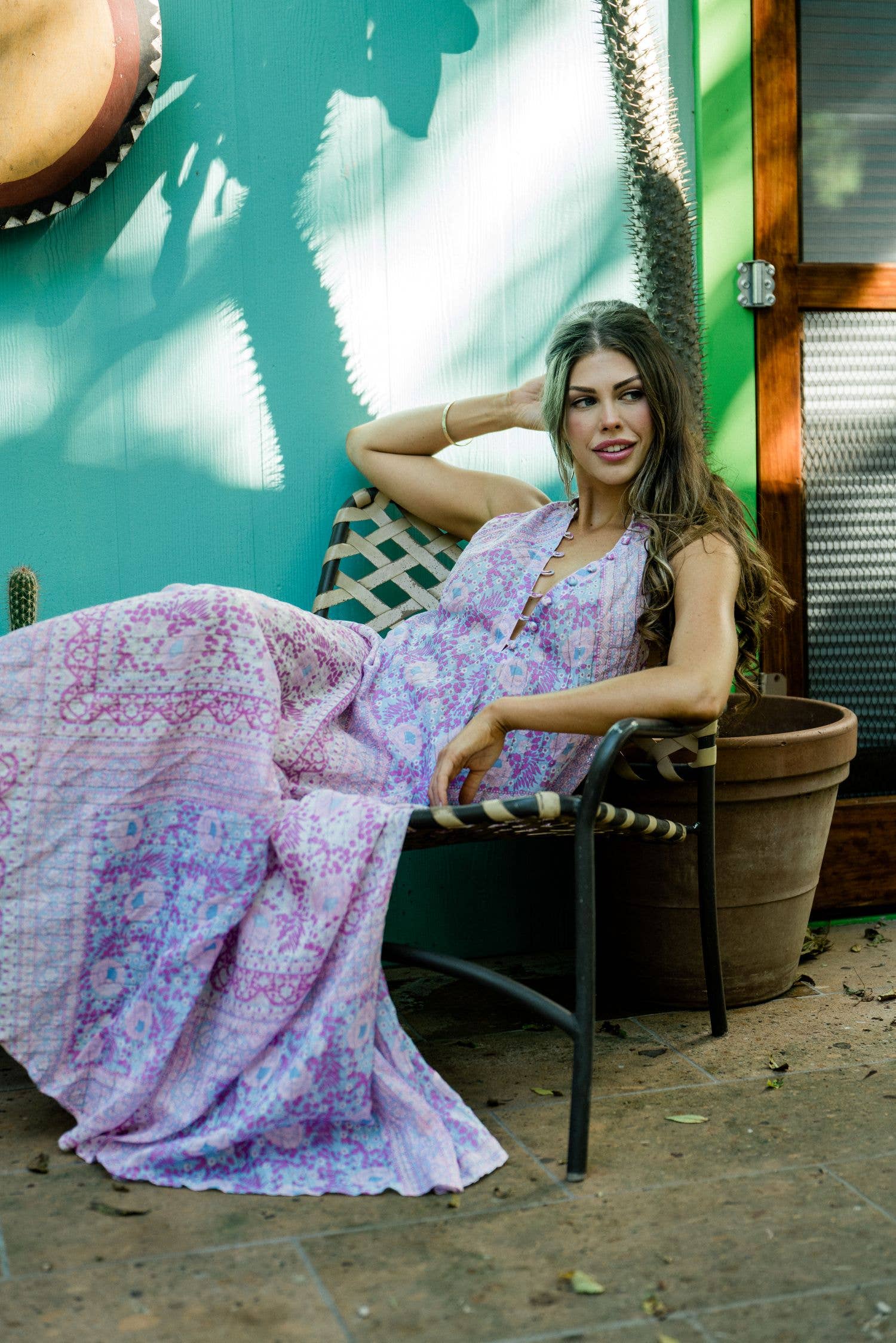 Woman relaxing in outdoor chair wearing a pastel pink and purple Cotton Glendora Maxi Dress in a sunlit garden setting