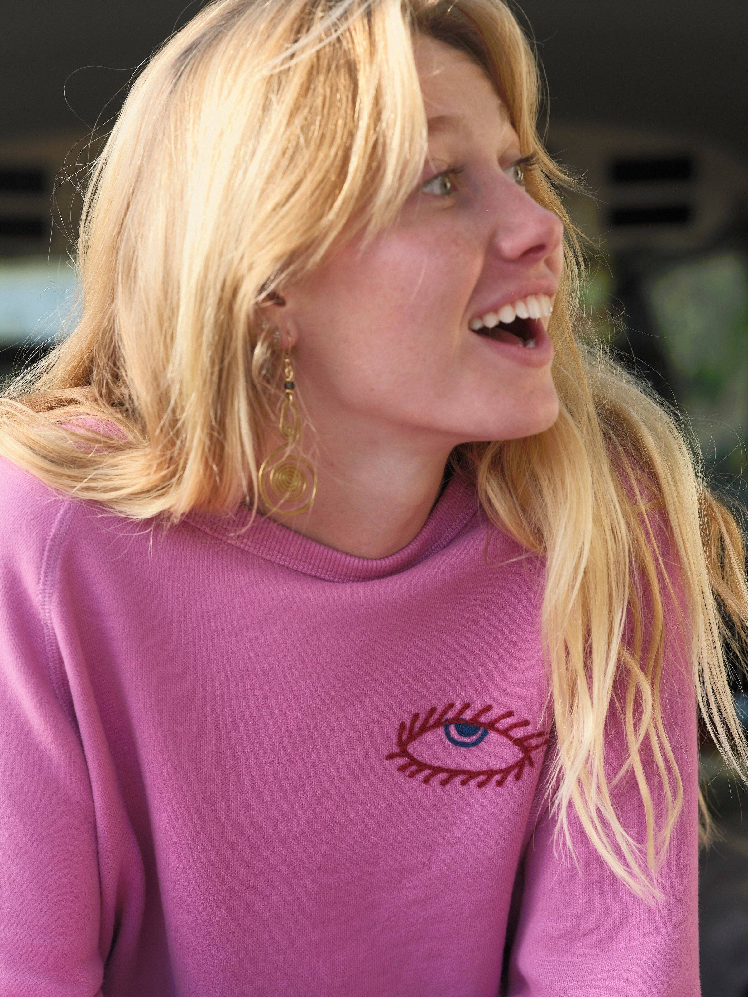 A woman with long blonde hair wears the Mollusk Visions Crew, a relaxed fit pink sweatshirt with an embroidered eye design. She smiles, accessorized with gold earrings, outdoors against a blurred background.