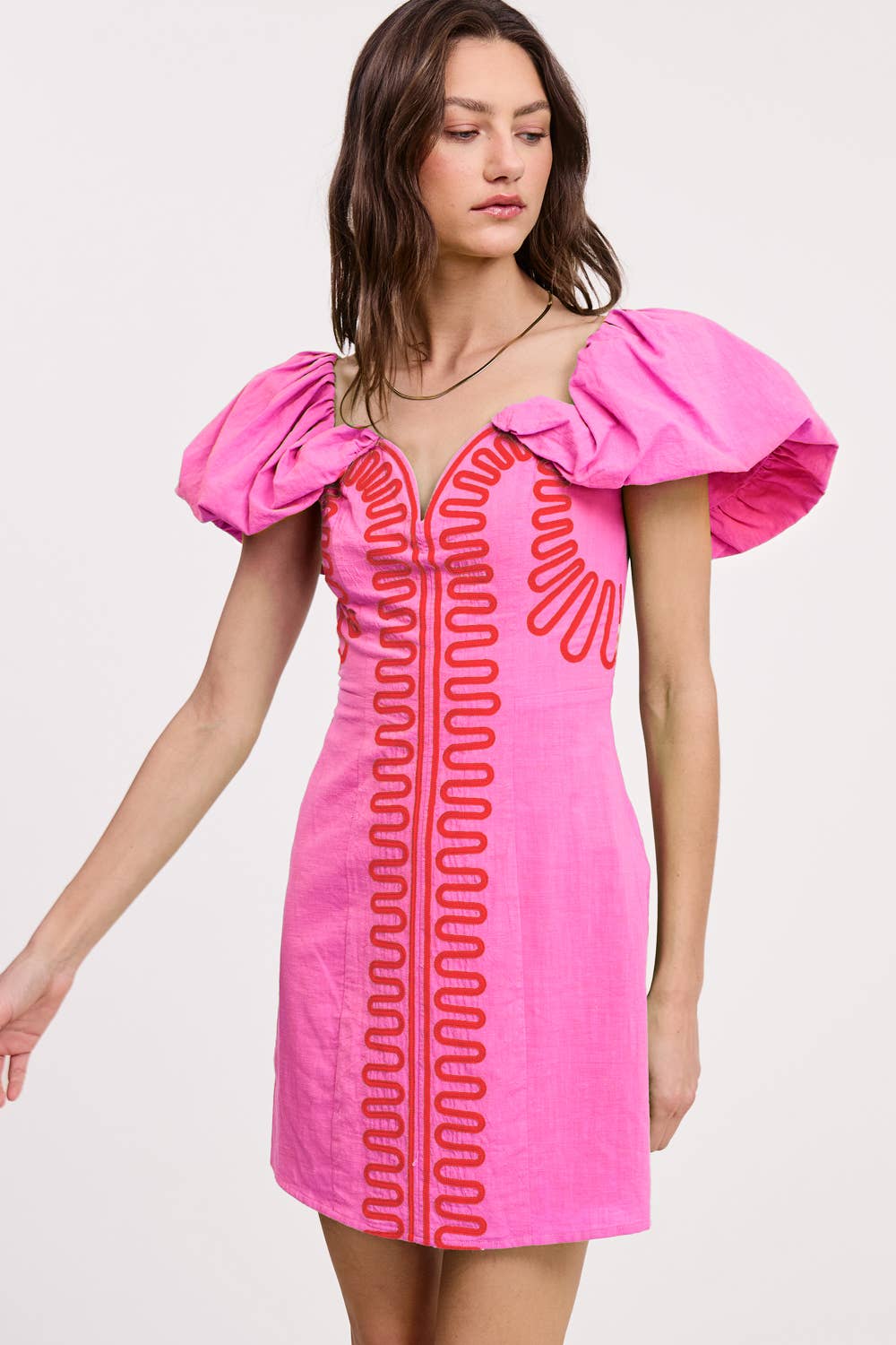 A woman models the Ultramarine Embroidery Detail Off Shoulder Linen Mini Dress, featuring bright pink fabric, bold puffed sleeves, and red embroidered trim. She poses against a plain light background, gazing to her left.