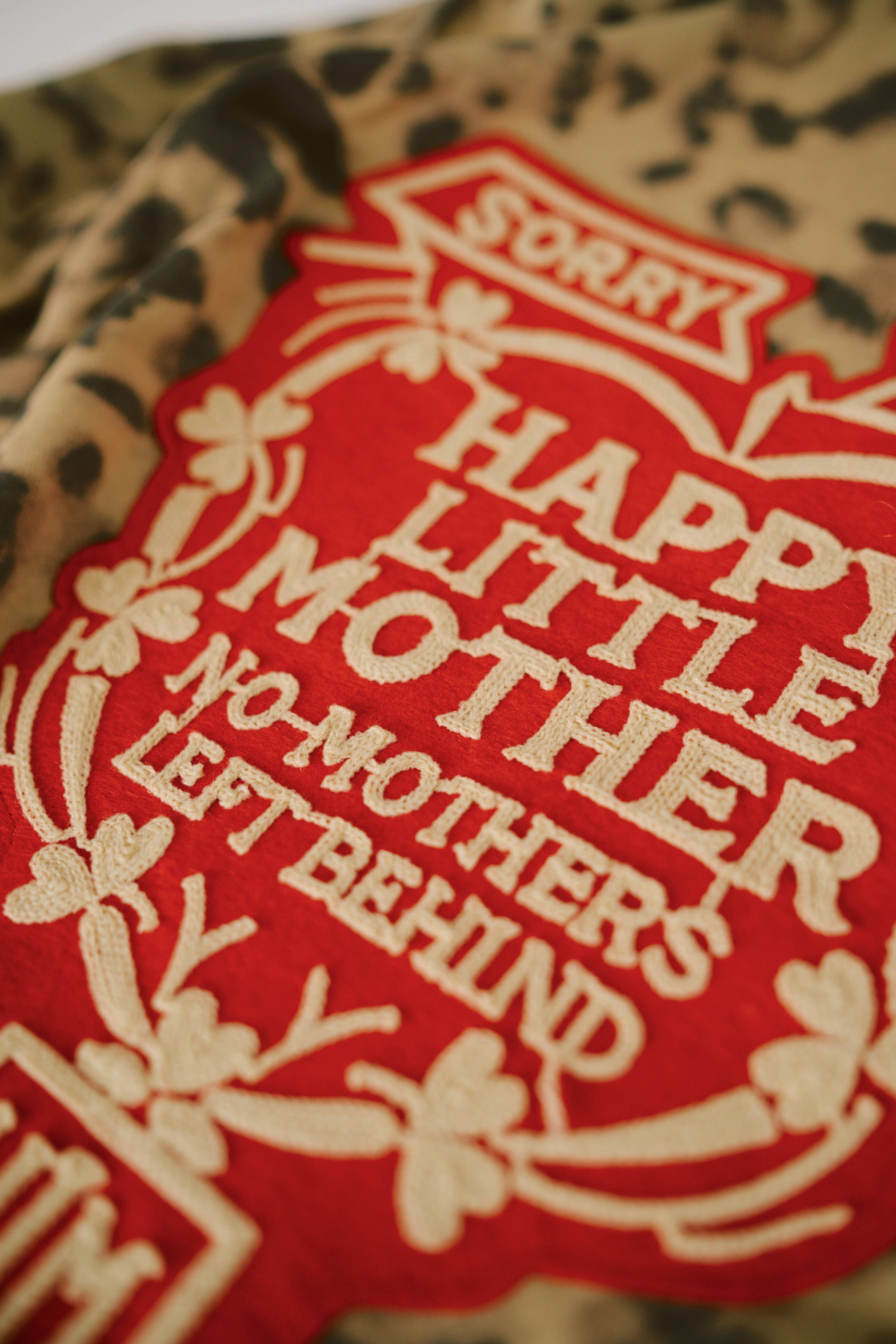 Close-up of embroidered patch with cream text on red background on SORRY MOM HOODIE - PRESALE with leopard print fabric.