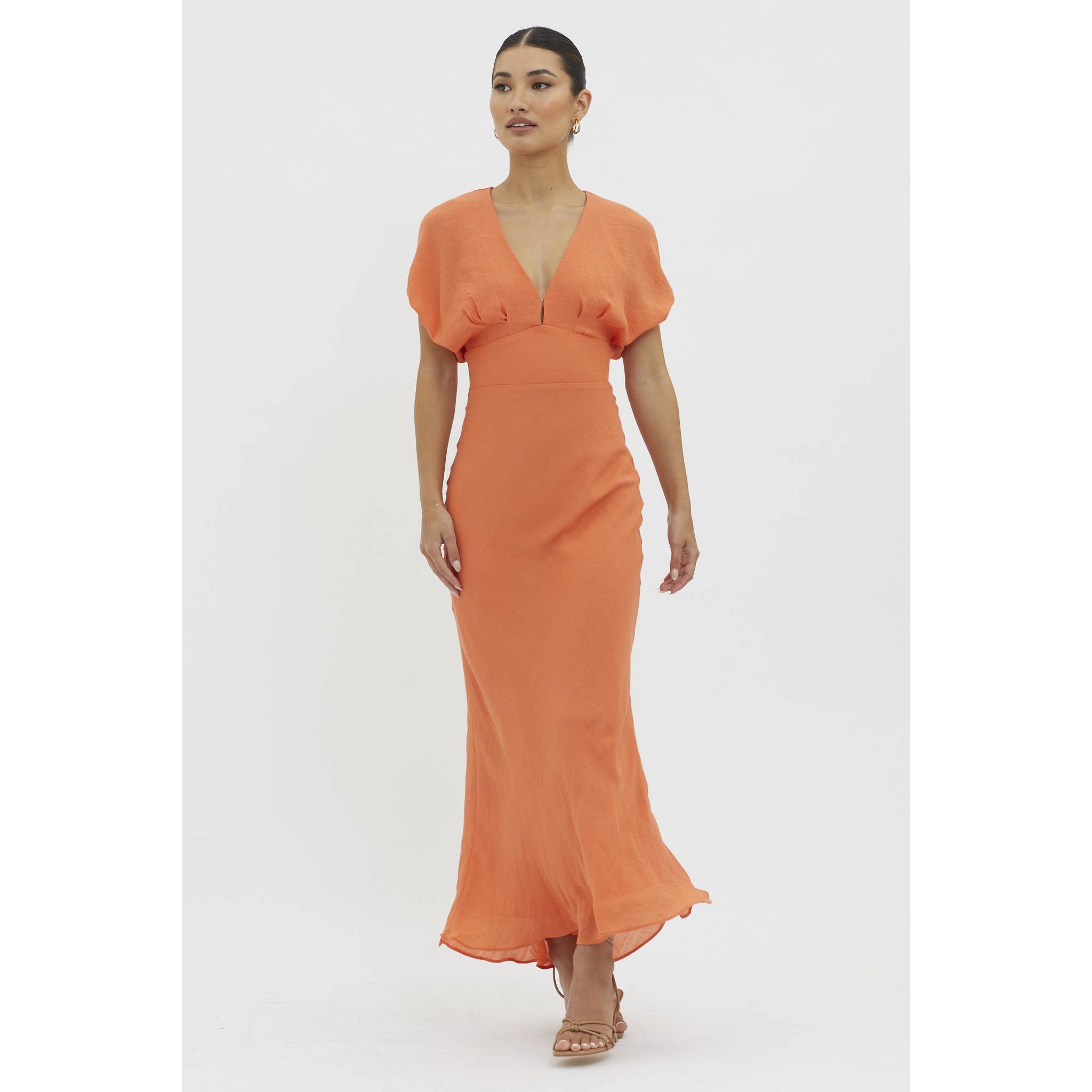 A woman with her hair pulled back and a neutral expression wears the One and Only Collective Tangerine V-neck Maxi Dress—an ankle-length, orange dress with an elastic waistband and short sleeves—paired with strappy sandals against a white background.
