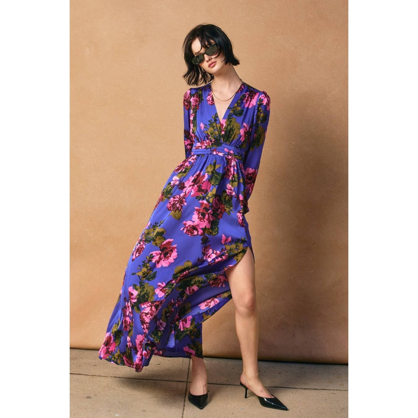 A person stands against a tan background, wearing the Dress Forum Mesmerizing Essence Pleated Waist Maxi Dress with a floral print and high side slit, styled with black pointed heels and dark sunglasses.