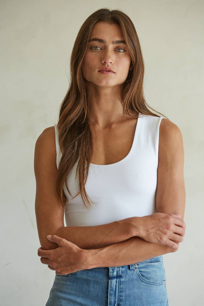A woman with long brown hair wears a white By Together Double Layered Seamless Top and blue jeans, standing against a plain light background. She looks directly at the camera with her arms loosely crossed.
