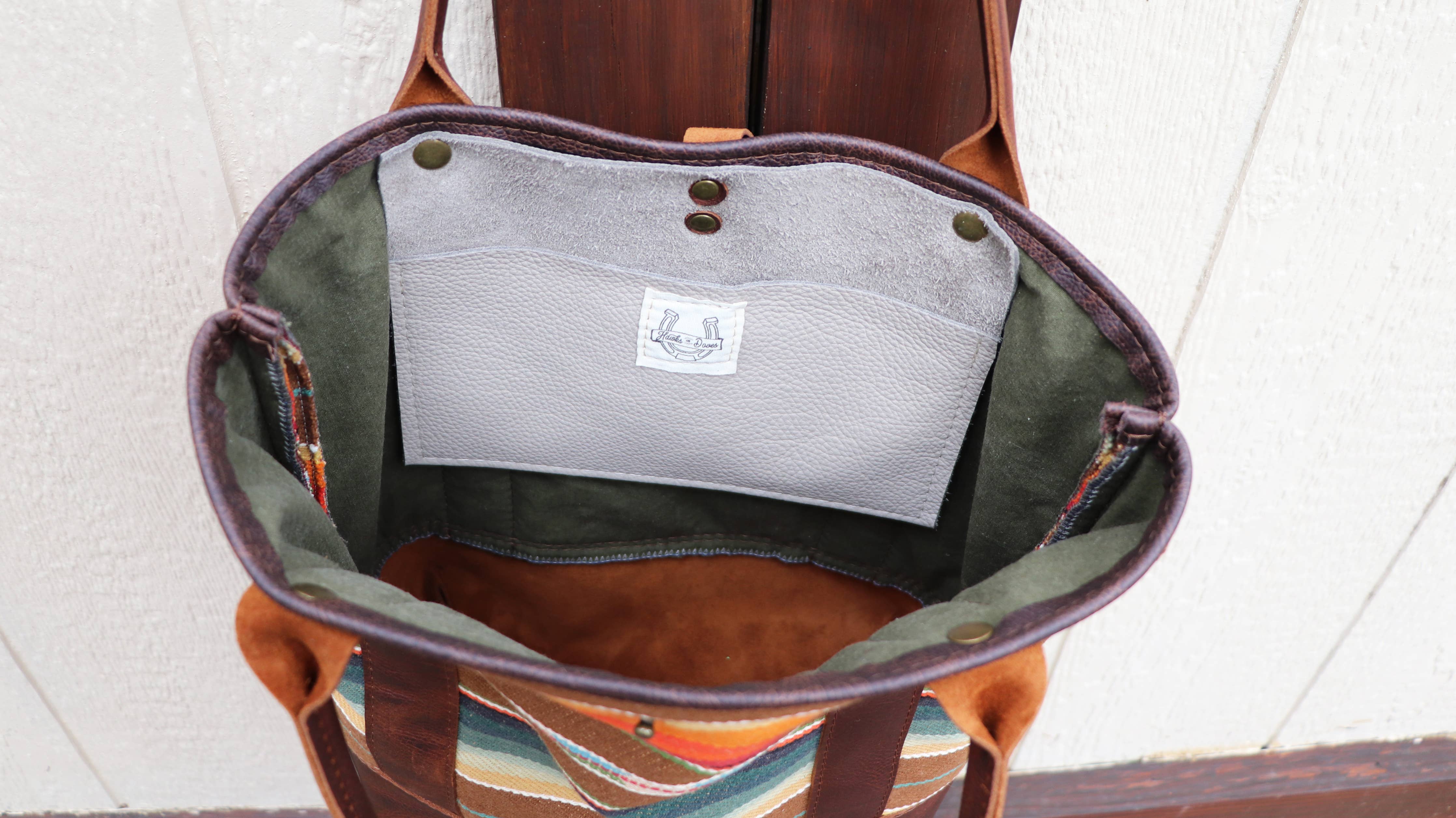 The Pearl Serape Tote by Hawks & Doves is a handmade open full grain leather tote with serape fabric sides, green interior trim, and a gray pocket featuring a white patch label with a boat illustration, shown upright against a wood and white wall.